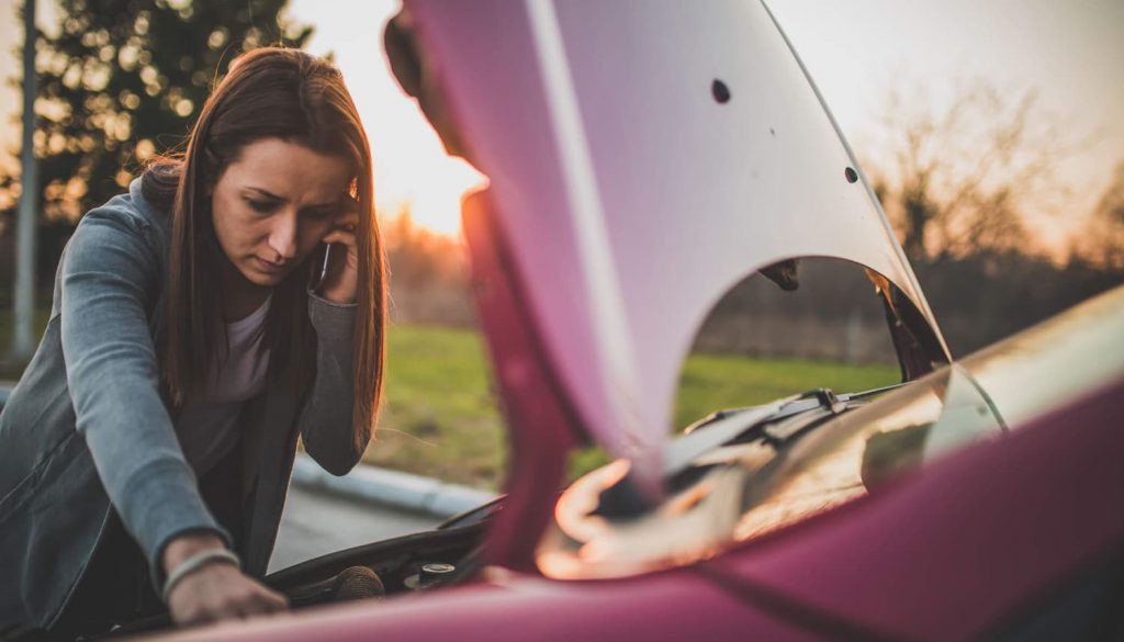 Mulher olhando algo no motor do carro e conversando com alguém no celular.